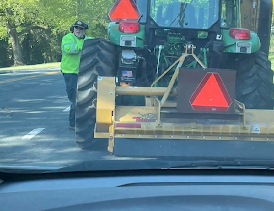 A worker wearing a bright green safety shirt and a cap stands beside a large green tractor on the road.