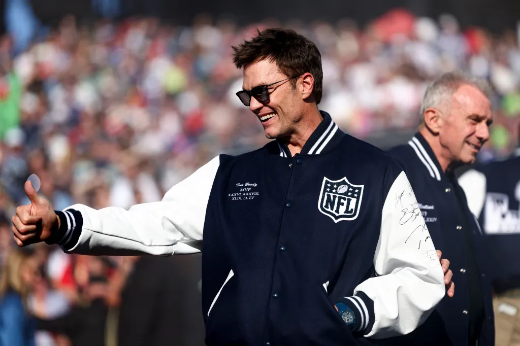Tom Brady smiling and giving a thumbs-up to fans in front of a stadium crowd.