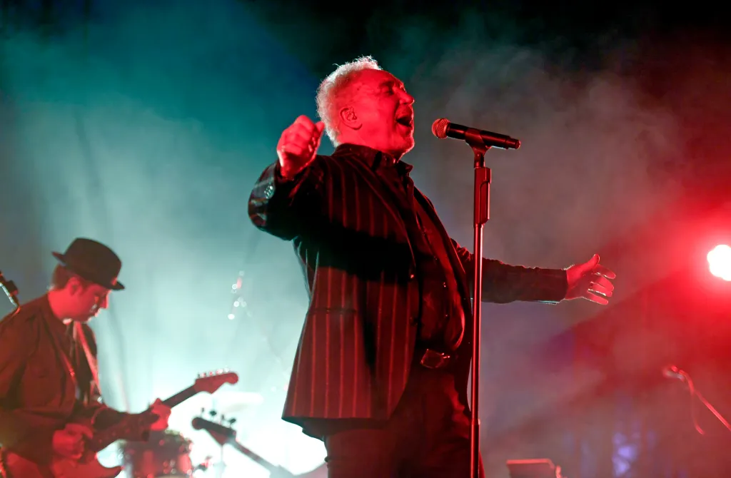 Tom Jones performs onstage during a music festival, singing into a microphone with dramatic red and green lighting.