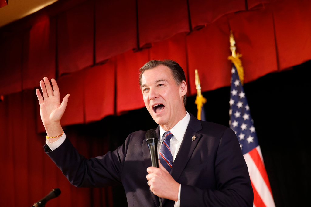 Tom Suozzi speaks into a microphone with his right hand raised at a get-out-the-vote rally.