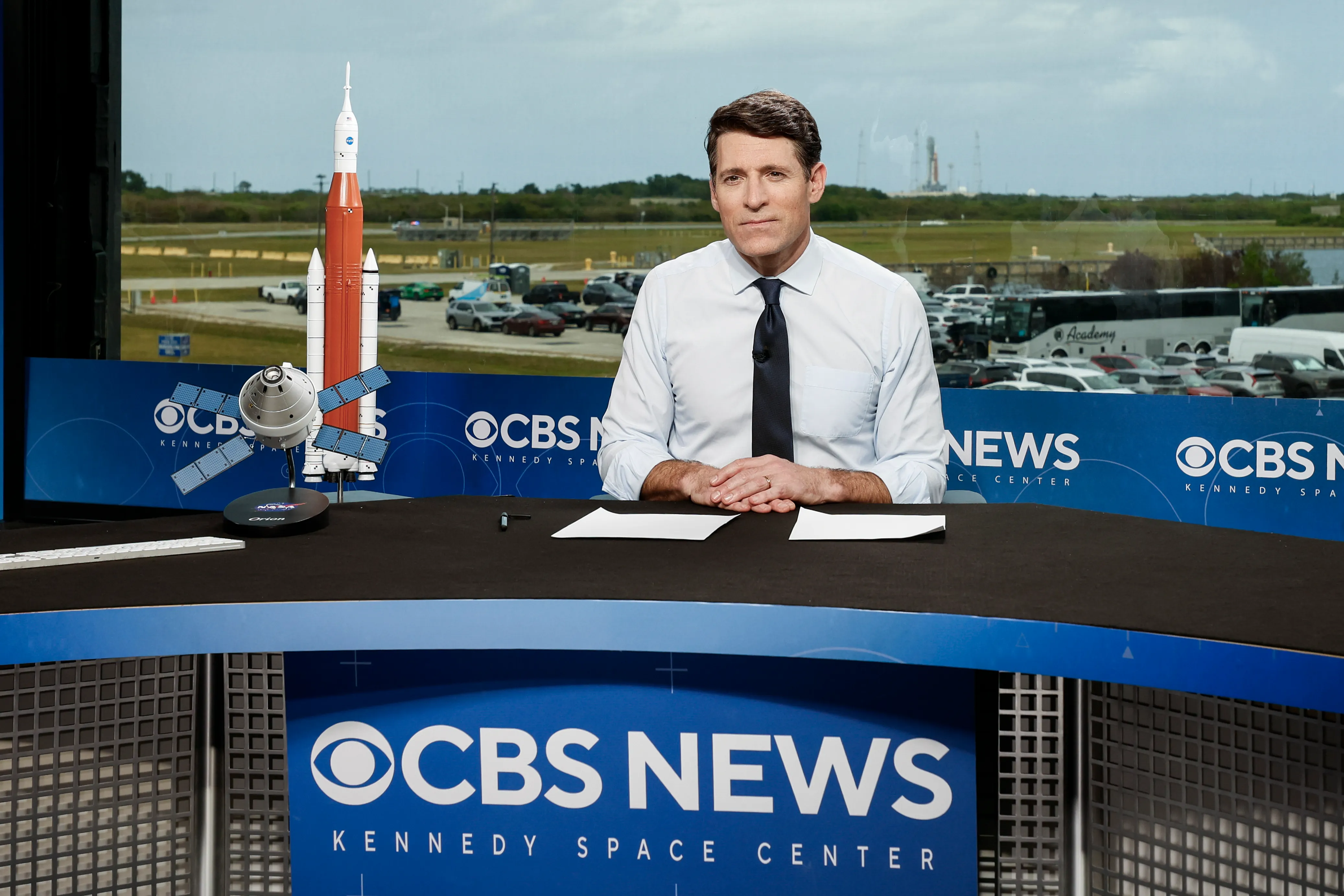 Tony Dokoupil anchors the CBS News broadcast from the Kennedy Space Center, with a model rocket on the desk and the launchpad visible through the window.