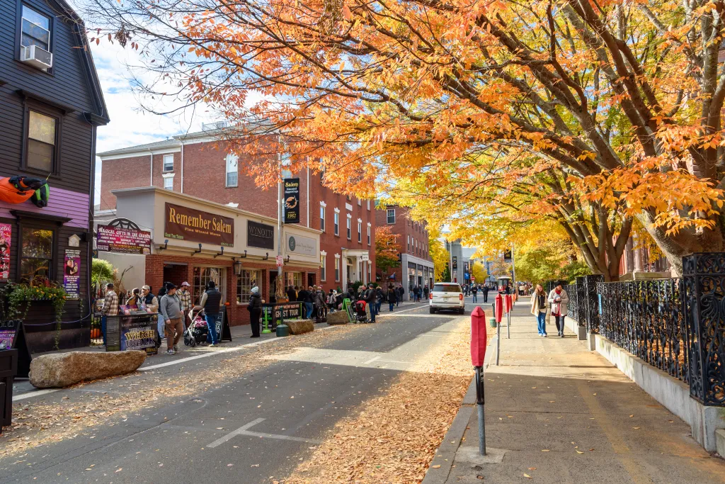 Tourists walk along a sunlit Essex Street in Salem, Massachusetts, framed by trees with vibrant autumn foliage.