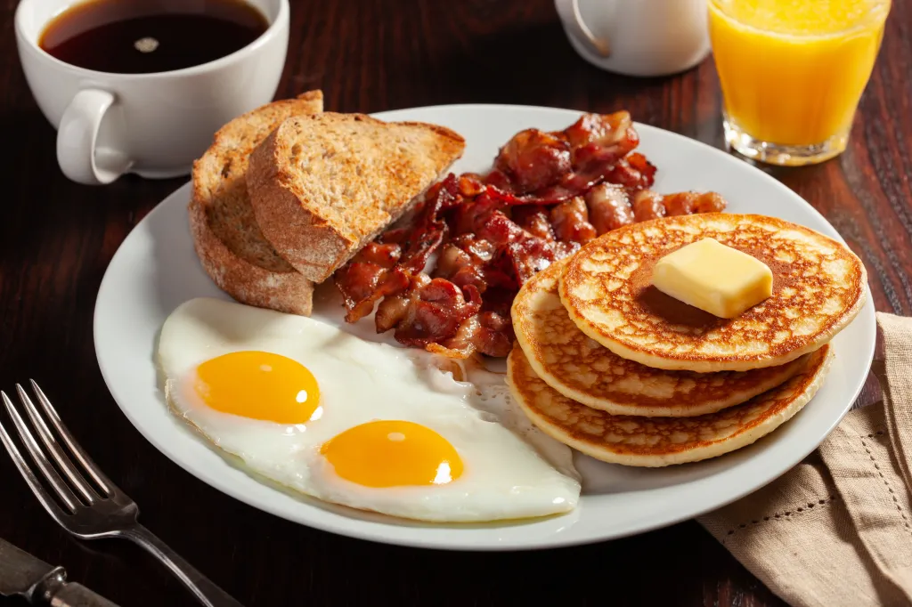 A traditional American breakfast featuring fried eggs, pancakes with butter, bacon, and toast, served on a plate with coffee and orange juice.