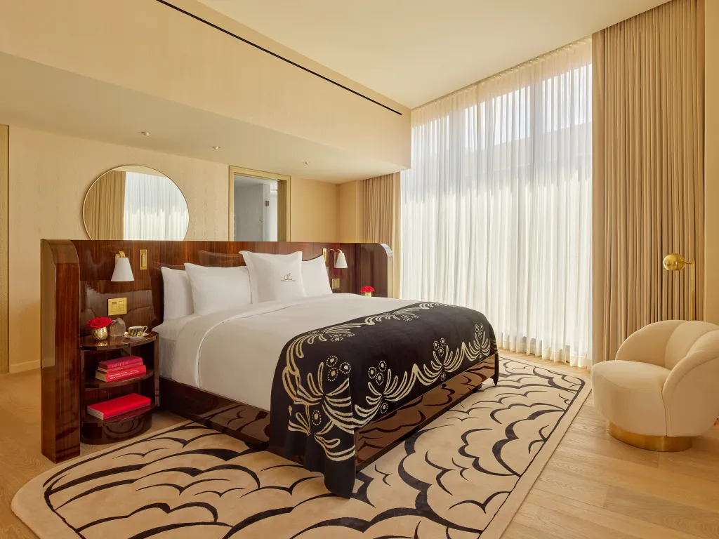 A hotel room with a large bed, a dark wood headboard, and floor-to-ceiling windows covered by sheer white curtains at Faena New York.