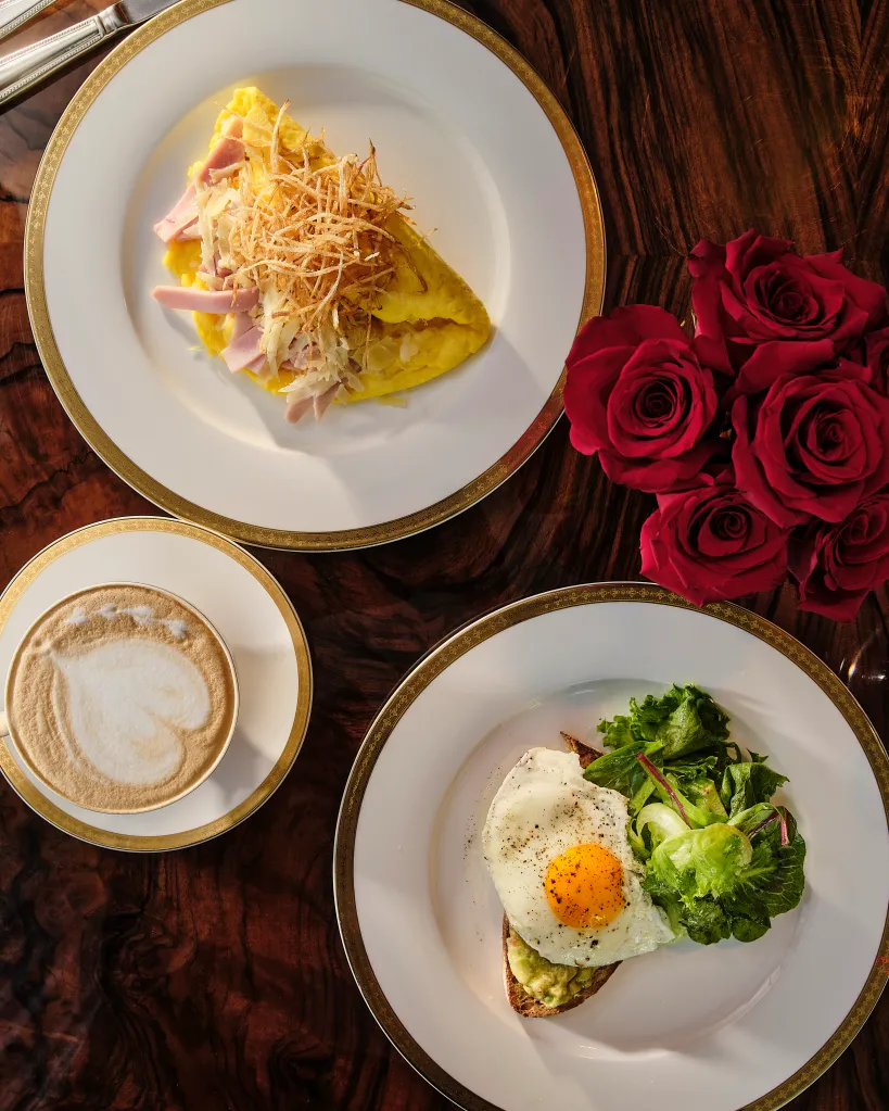 An overhead view of a breakfast spread featuring an omelet with ham and shoestring potatoes, a fried egg on avocado toast with side salad, a latte with heart-shaped latte art, and a bouquet of red roses on a wooden table.