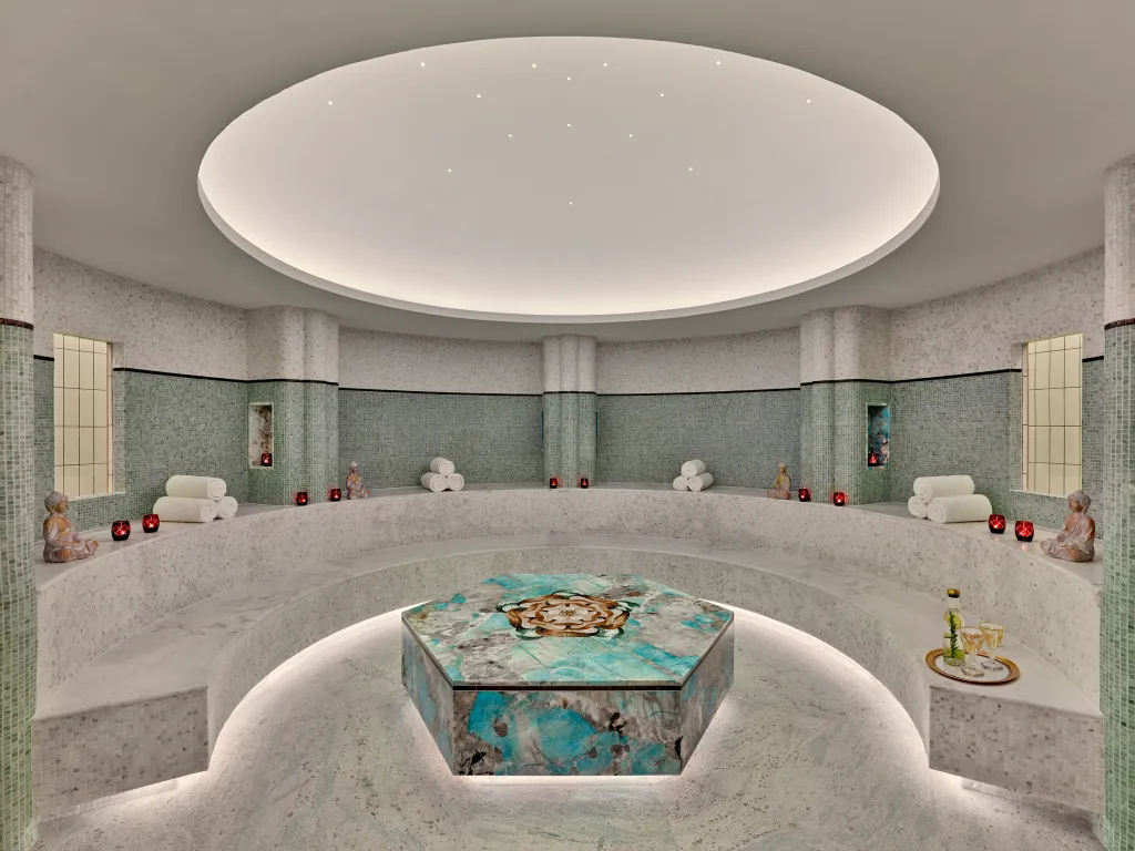 A circular tiled steam room with a central marble platform, curved marble seating, rolled towels, and lit candles at Faena New York.