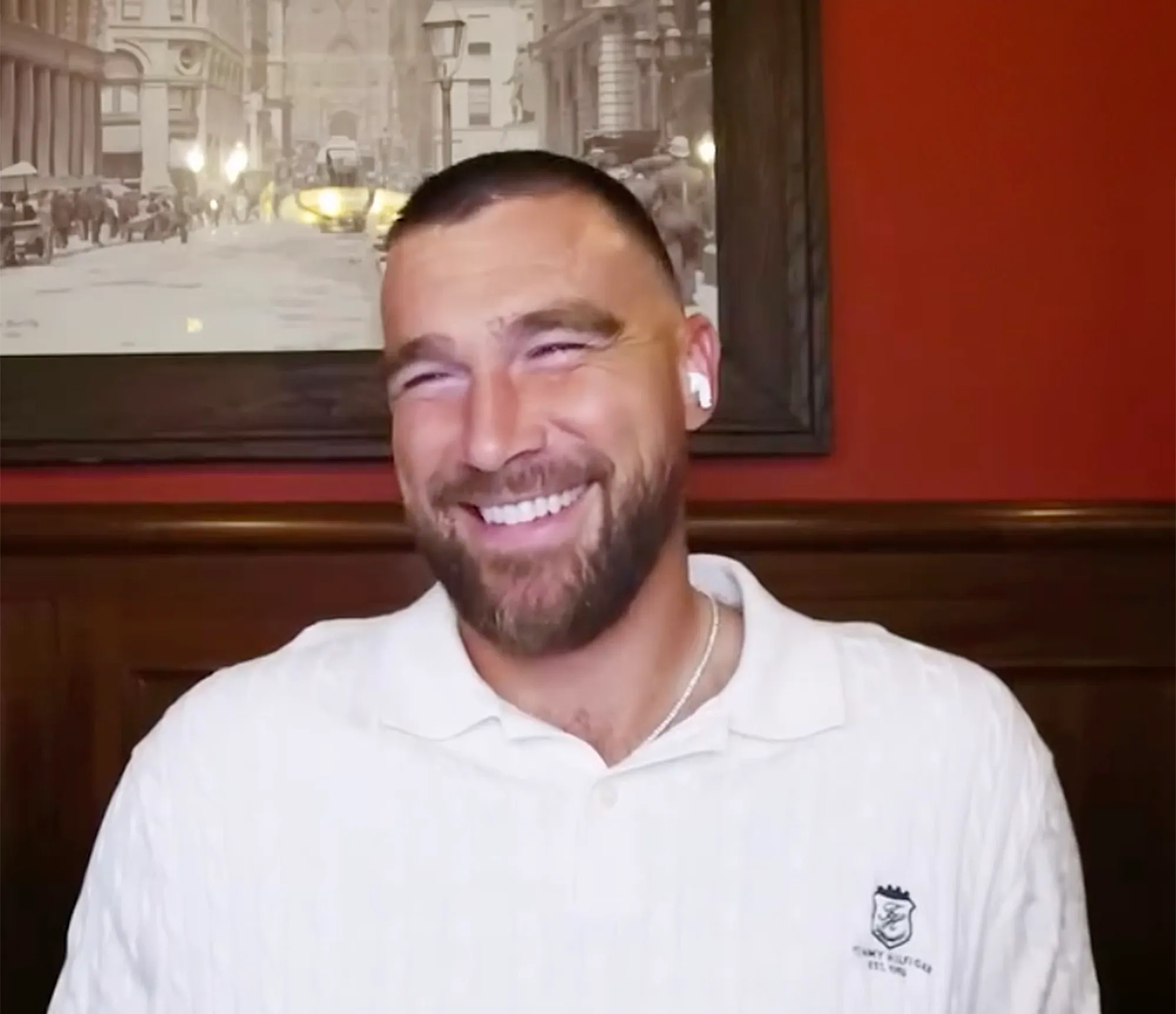 Travis Kelce smiling in a white polo shirt with a framed picture in the background.