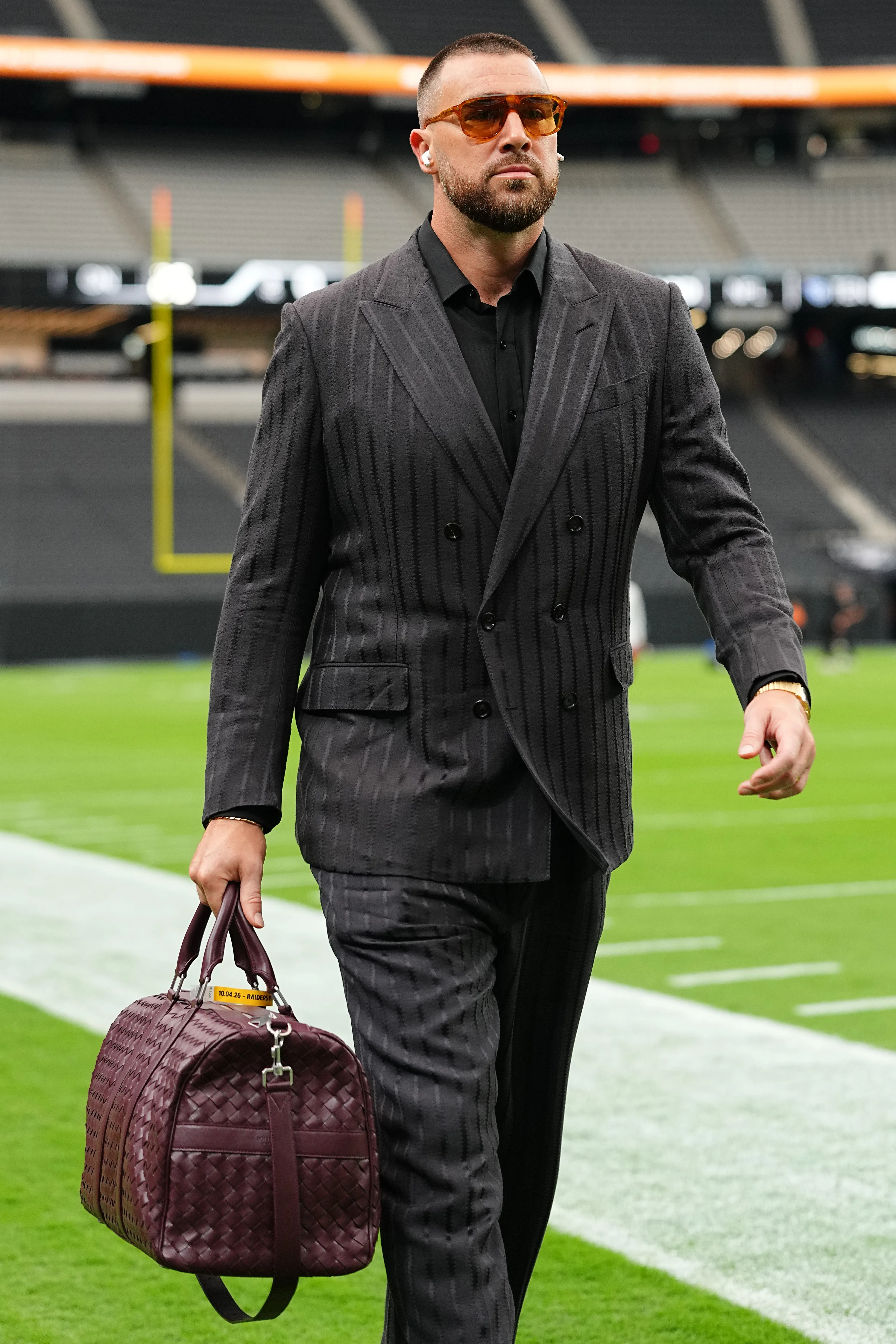 Travis Kelce of the Kansas City Chiefs arrives at Allegiant Stadium in a patterned gray suit and sunglasses, carrying a maroon duffel bag.