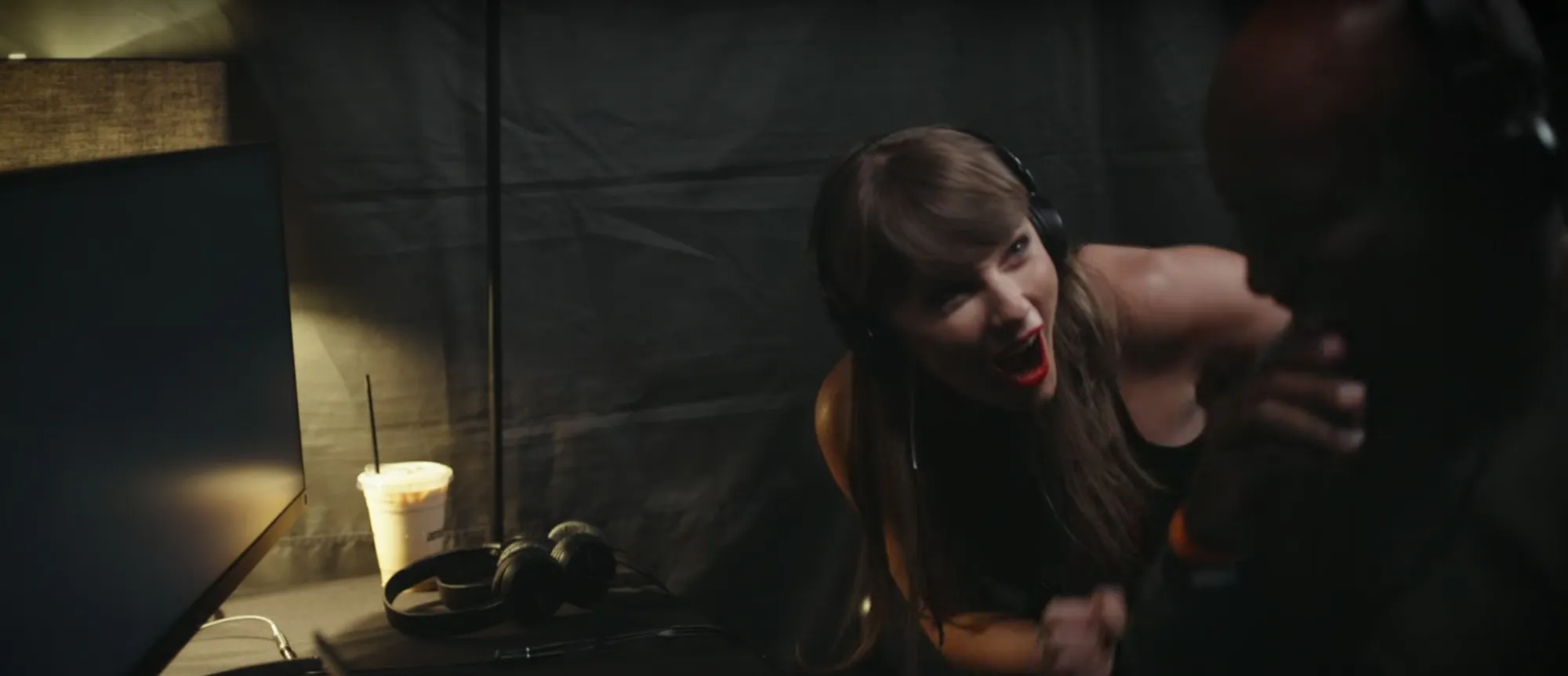 Taylor Swift wearing headphones and smiling while leaning in towards Travis Kelce in a behind-the-scenes shot.