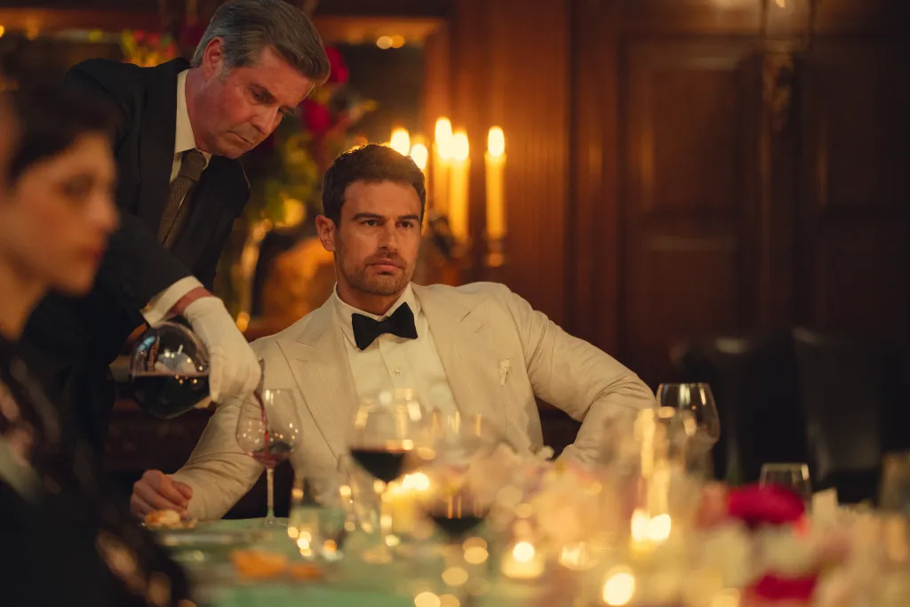 Theo James seated at a dining table while a butler pours wine.