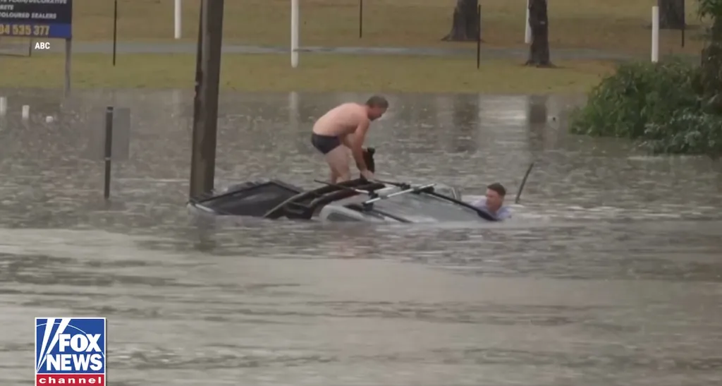 A rescuer and a TV reporter help a man trapped in a sinking vehicle during floodwaters.
