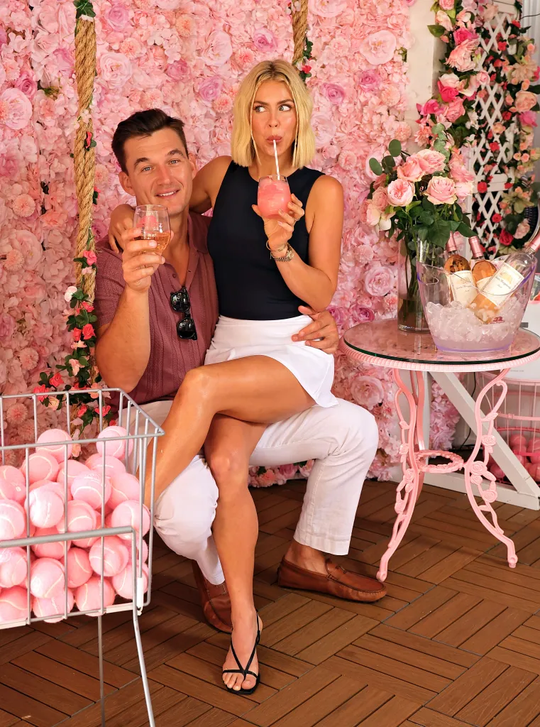 Tyler Cameron and Tate Madden pose together while holding drinks in front of a pink flower wall.
