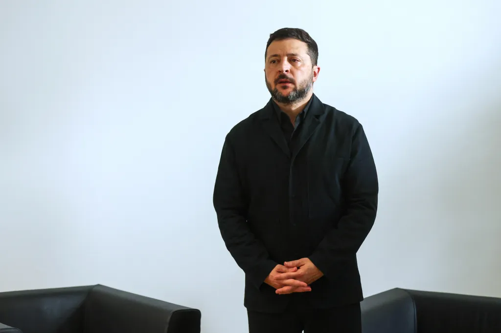 Ukraine's President Volodymyr Zelenskyy stands in a room with white walls, clasping his hands in front of him.