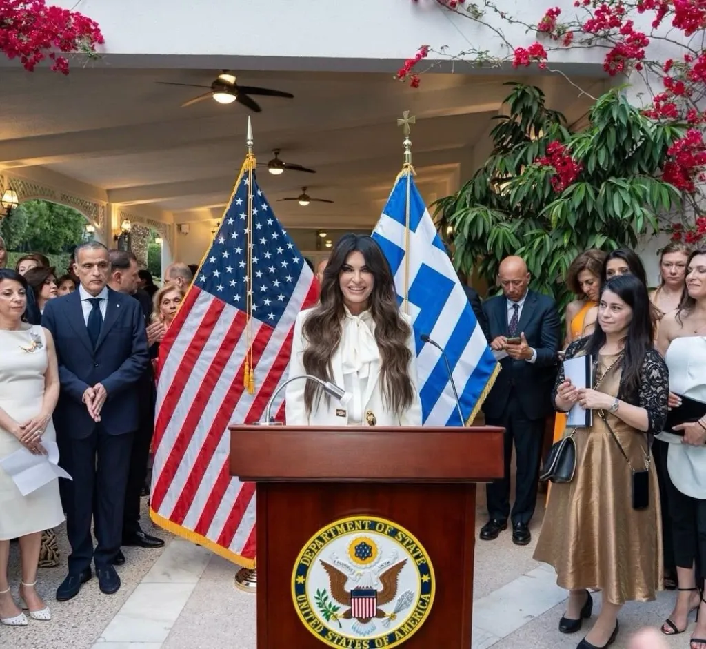 US Ambassador to Greece Kimberly Guilfoyle stands at a podium with US and Greek flags behind her, surrounded by other people.