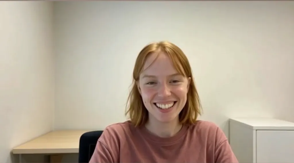 An AI-generated video agent created by Tavus, depicting a smiling woman with reddish-blonde hair in an office setting.