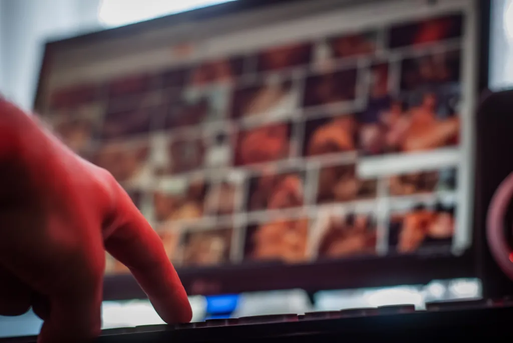 A person's hand reaches toward a computer keyboard while a monitor displays adult content.