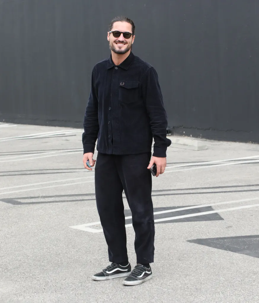 Val Chmerkovskiy standing in a parking lot, smiling and wearing a dark corduroy outfit and sunglasses.