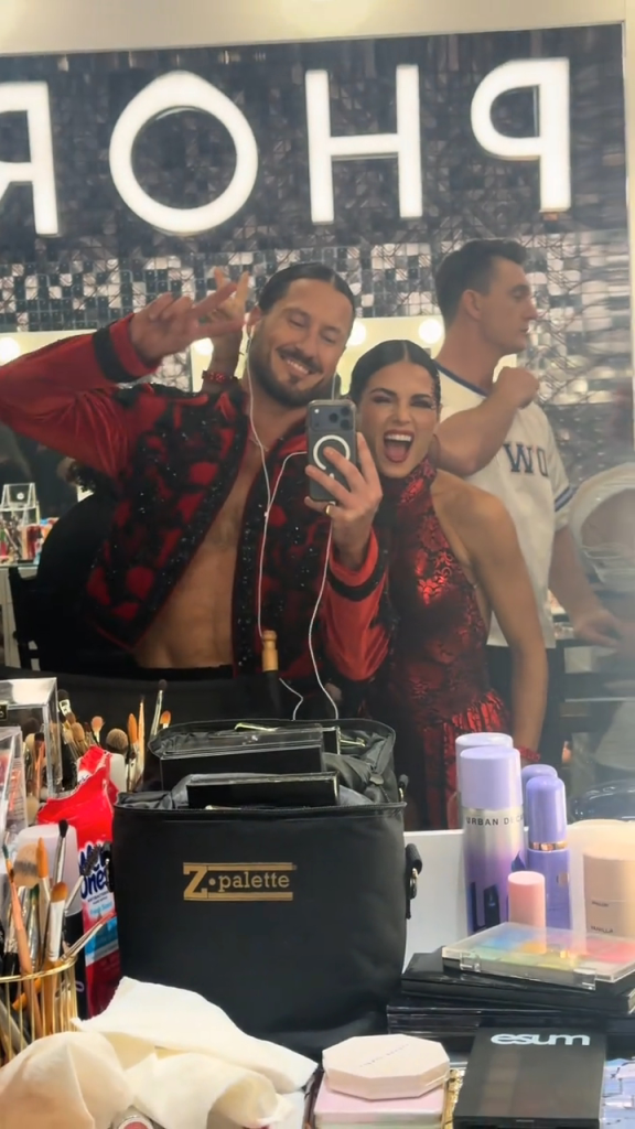 Val Chmerkovskiy and Jenna Johnson take a mirror selfie in a dressing room, with a man standing in the background.
