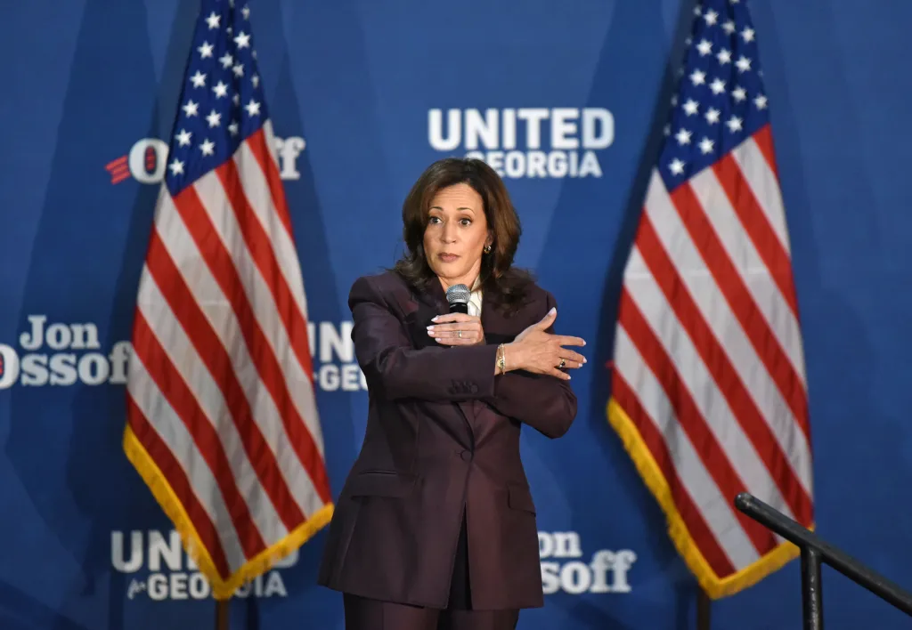 Vice President Kamala Harris speaks into a microphone while standing between two American flags at an event in Atlanta.