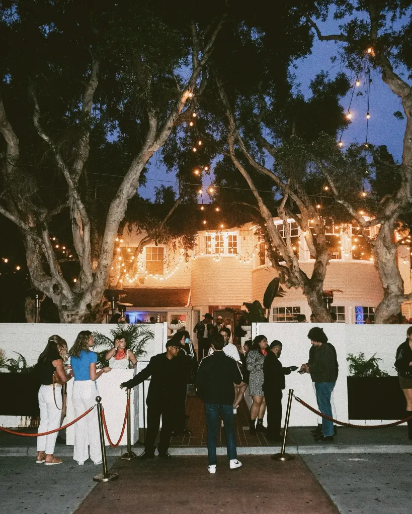 People gather outside The Victorian in Santa Monica, California, illuminated by string lights hanging from large trees.