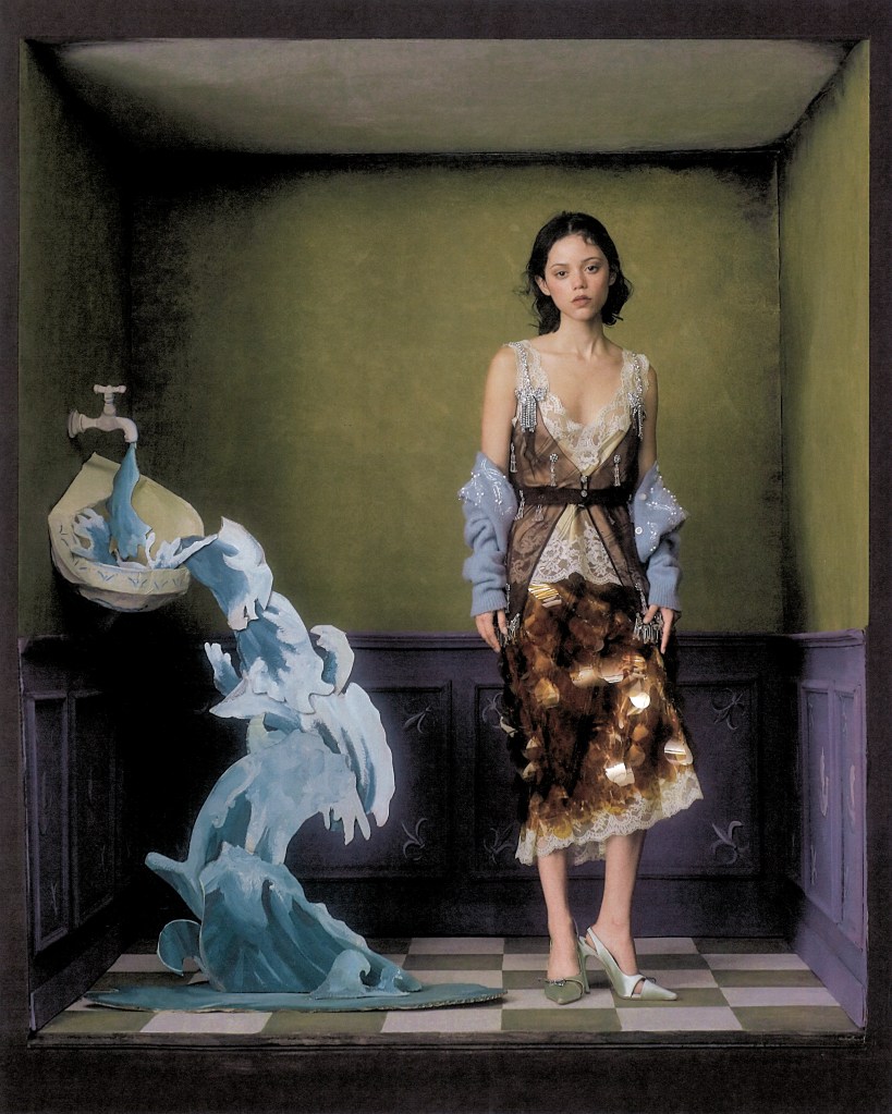 Jenna Ortega stands in a room with a checkered floor, next to a sculptural water feature flowing from a wall-mounted basin.