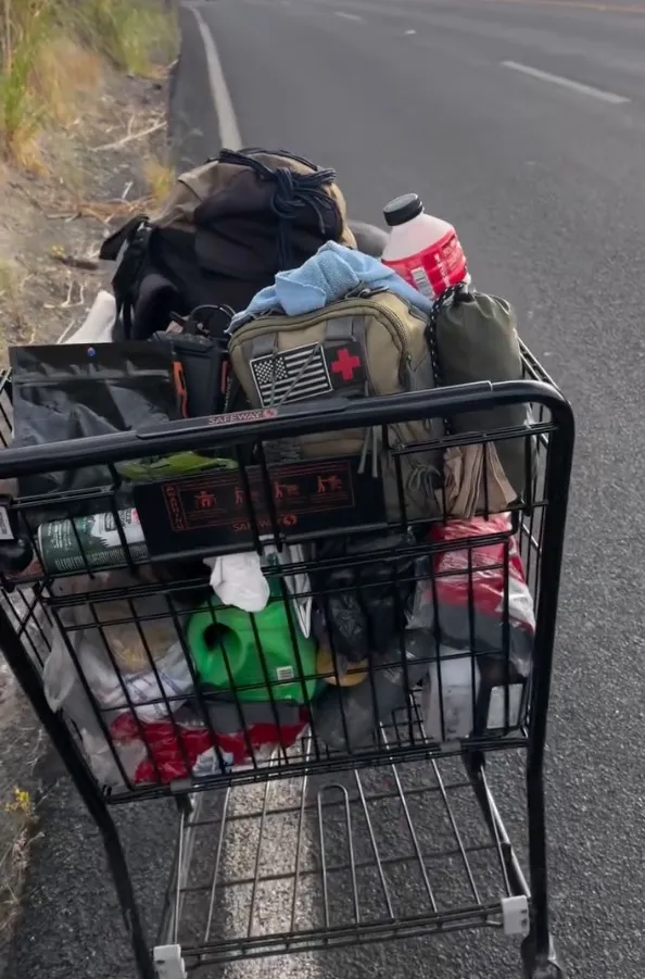 Chantz Mula's shopping cart loaded with supplies, including a backpack, water, and gear, as he walks along a roadside during his cross-country journey.