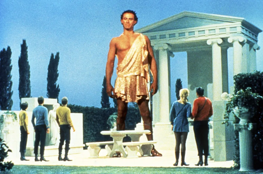 A scene from Star Trek featuring Walter Koenig, DeForest Kelley, William Shatner, Michael Forest as a giant, Leslie Parrish, and James Doohan in an outdoor setting with a classical temple.