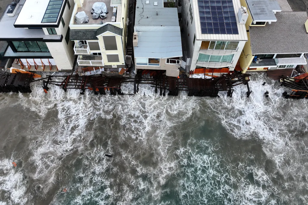An overhead view of high waves crashing against the foundations and decks of beachfront homes.