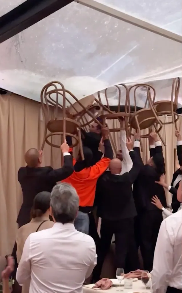 Wedding guests hold up chairs to support a sagging tent roof weighed down by rainwater.
