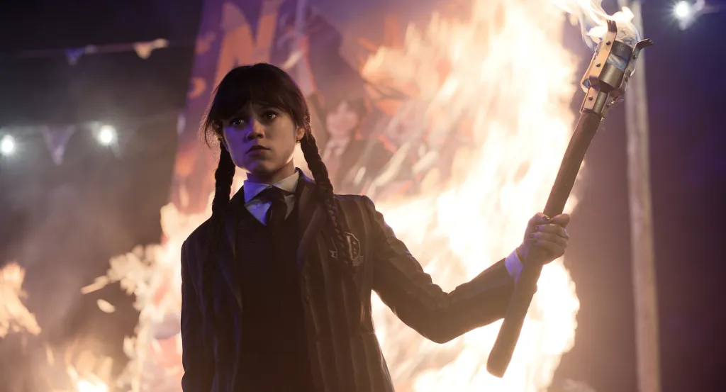 Jenna Ortega as Wednesday Addams holds a flaming torch in front of a fire.