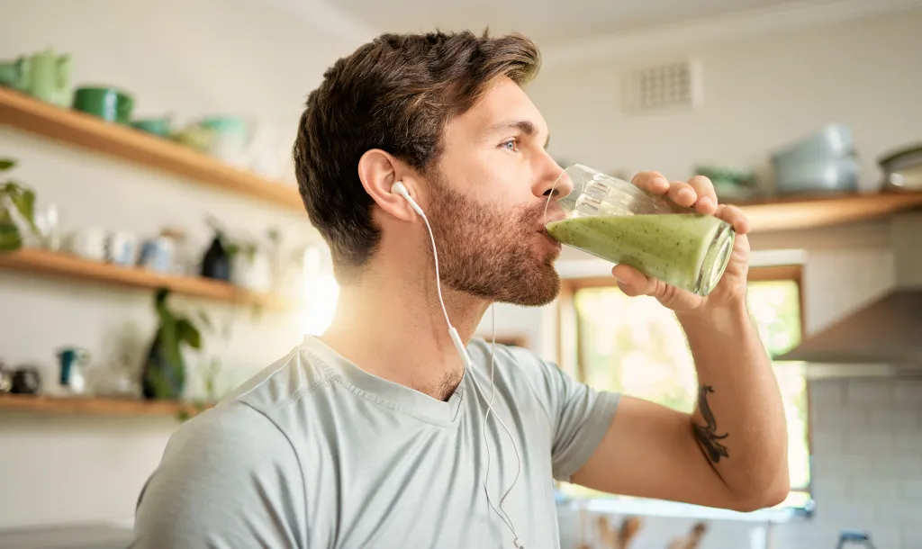 A man wearing earphones drinks a green smoothie in his kitchen.