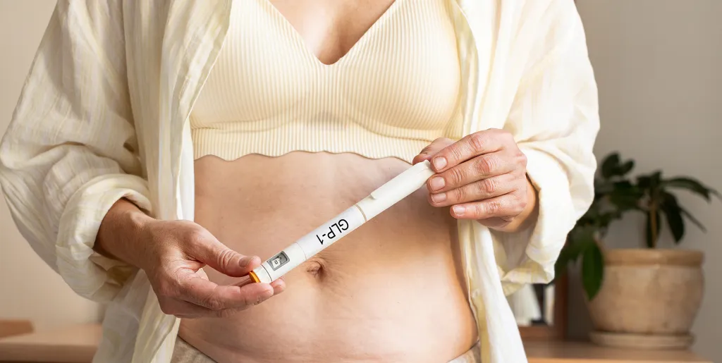 A woman holding a GLP-1 pen injector near her abdomen at home.