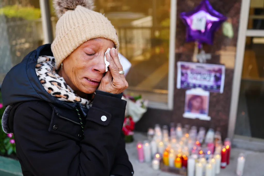 A woman in a winter hat cries beside a memorial of candles and a purple balloon.