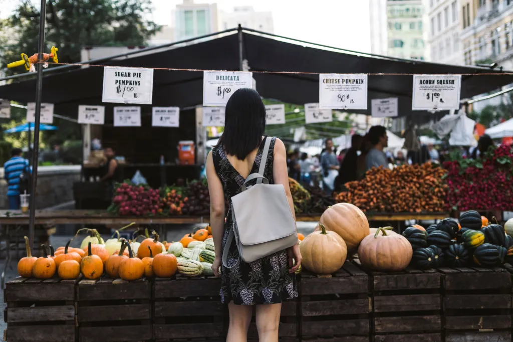 A woman with a backpack views produce at an outdoor street market.