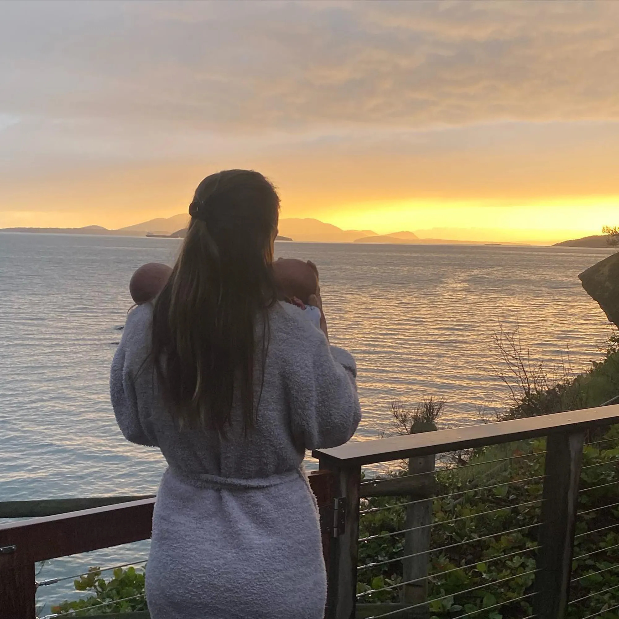 A woman holding twin babies looks out at the ocean at sunset.