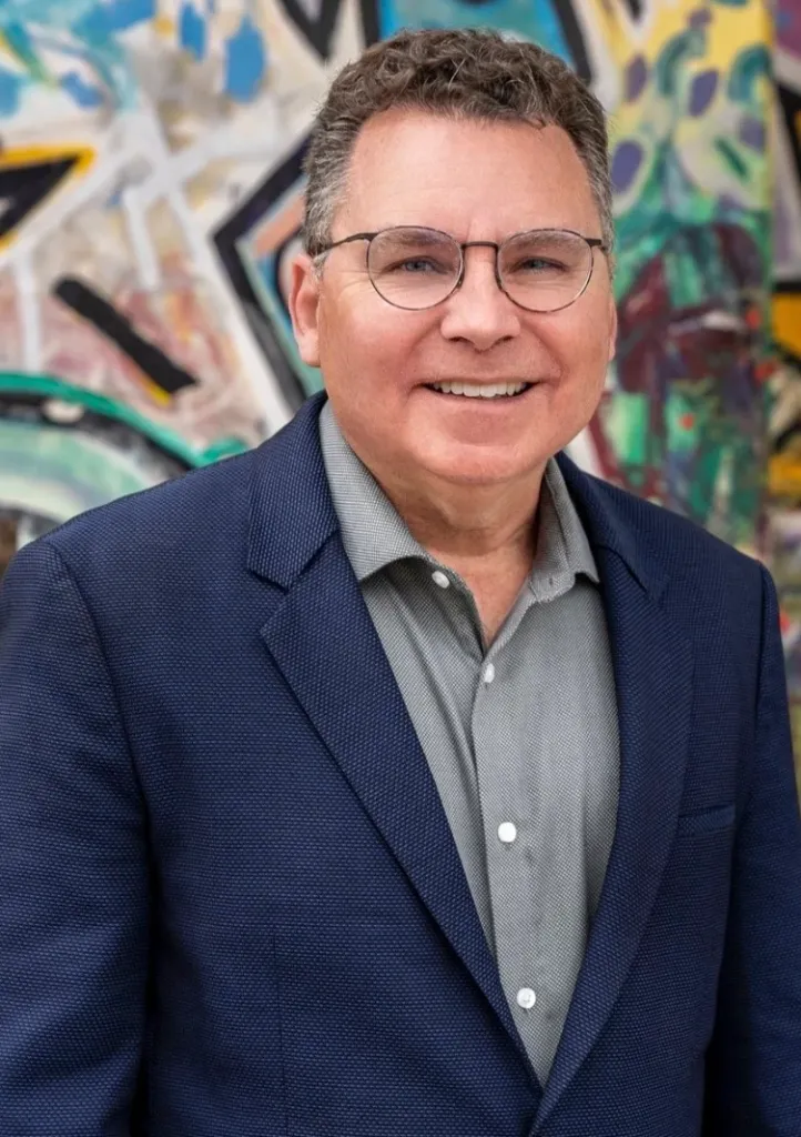 Richard Barrera smiling while wearing a blue blazer and glasses, with a colorful abstract background.