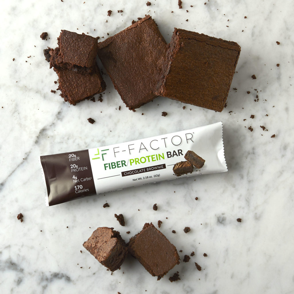 An F-Factor Chocolate Brownie Fiber/Protein Bar with chocolate brownie pieces and crumbs scattered on a white marble surface.