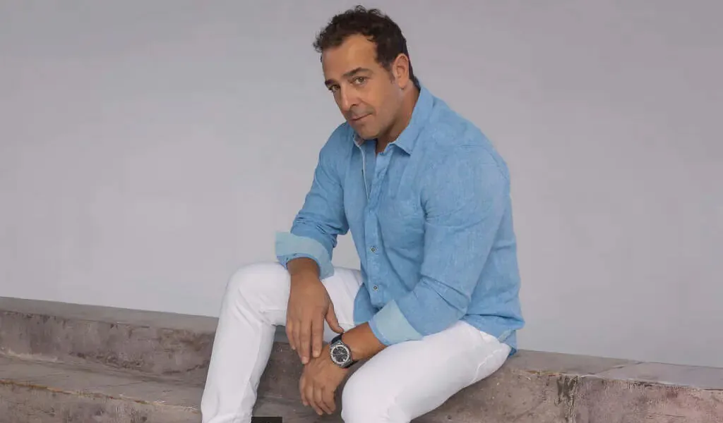 Portrait of Peter Mangiameli sitting on a concrete ledge, wearing a light blue button-down shirt and white pants.