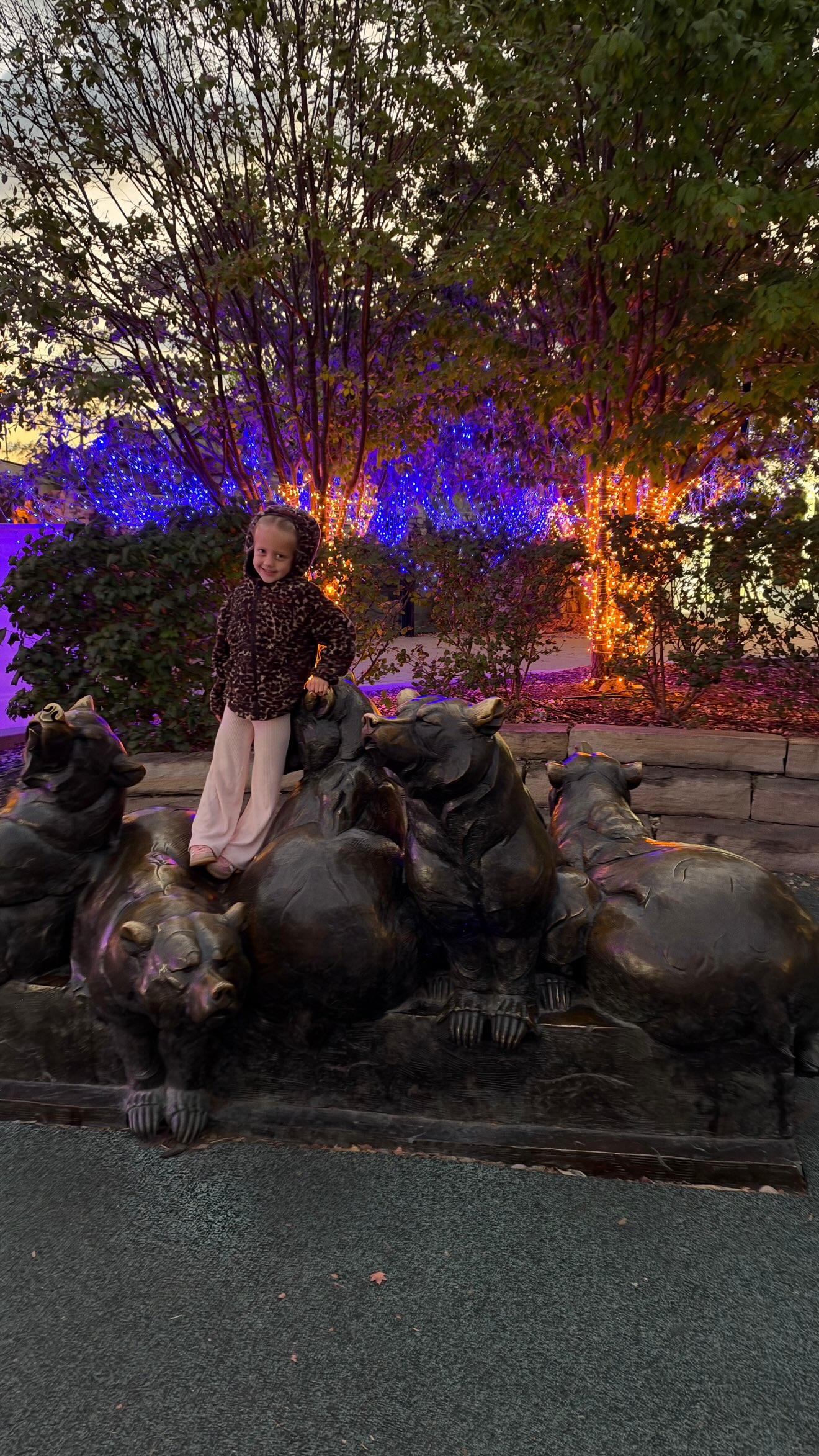 A young girl posing with bronze bear statues at night, with illuminated trees and colorful lights in the background.