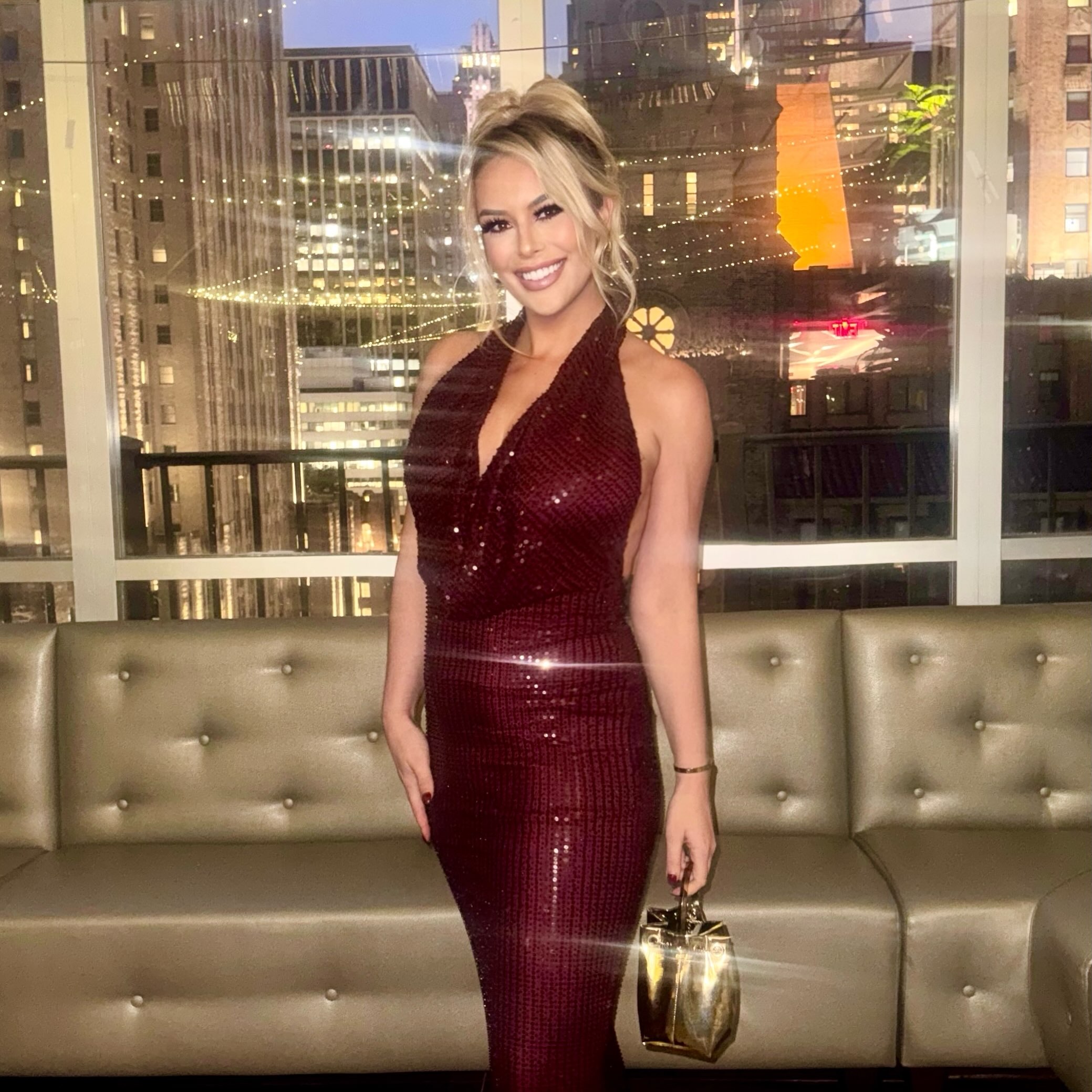 Britt McHenry in a sparkly maroon halter dress standing in front of a window and a couch.