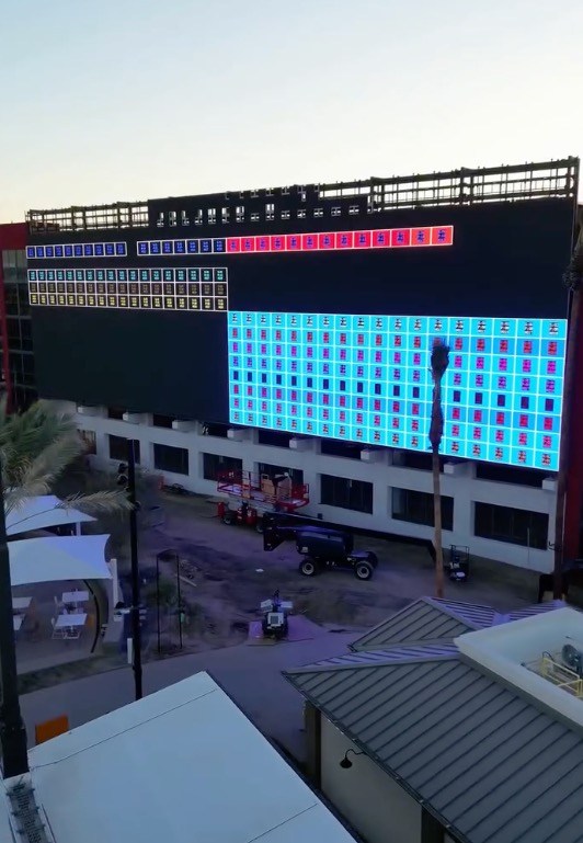 An exterior view of a large building under construction with a massive LED screen displaying grids of colored lights.