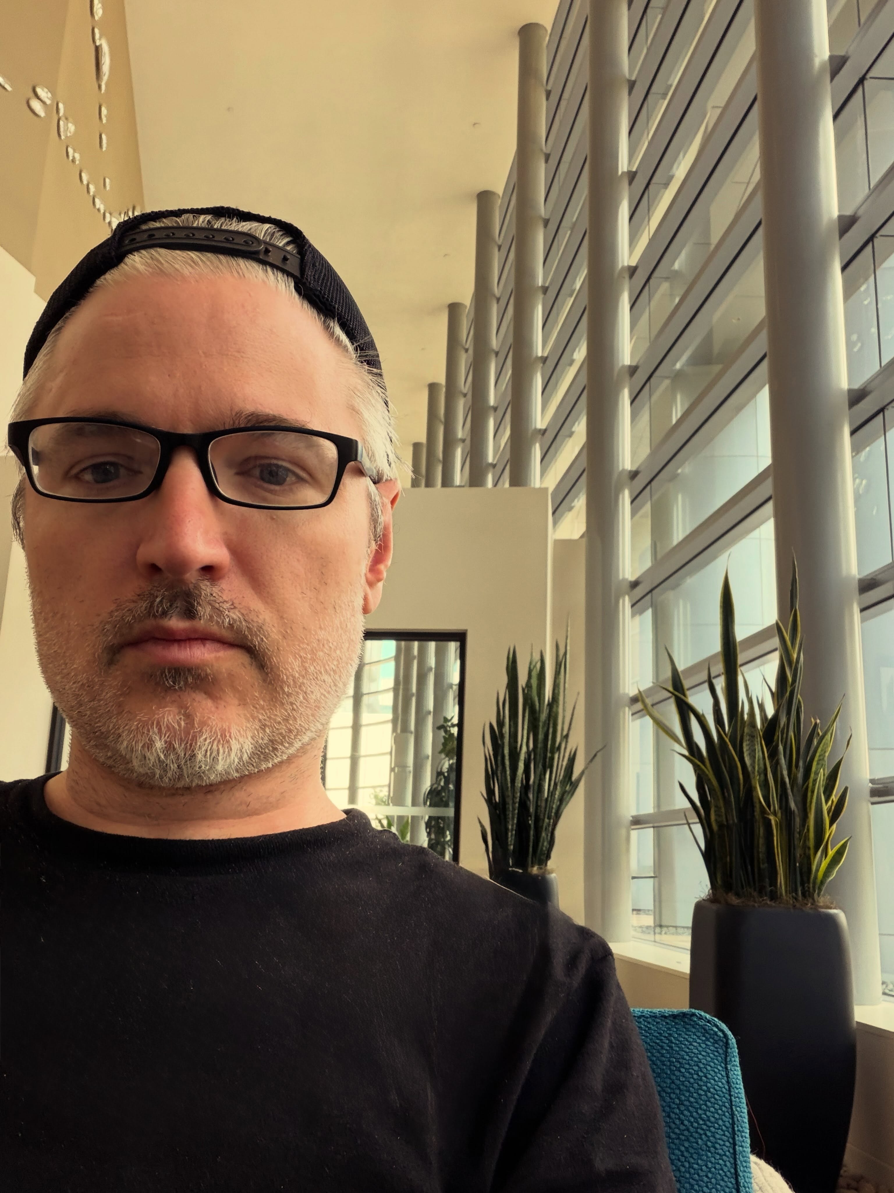 A close-up selfie of Lucian Piane wearing a black shirt, glasses, and a backward cap in a large room with tall windows and potted plants.