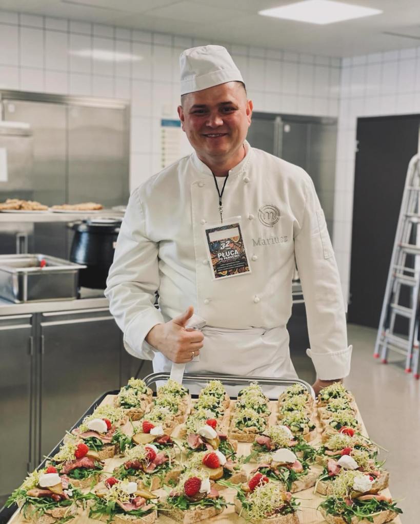 Polish 'MasterChef' star Mariusz smiling in a kitchen with a tray of open-faced sandwiches.