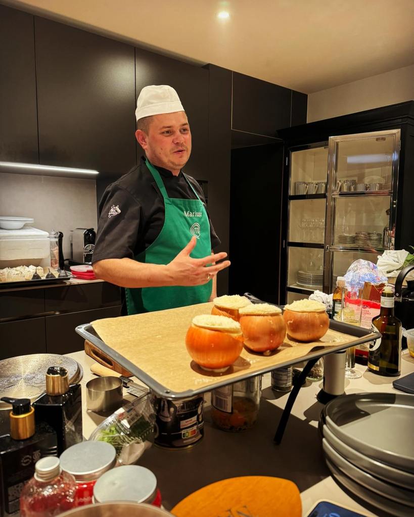 Chef Mariusz stands in a kitchen, wearing a chef's hat, black uniform, and green apron, gesturing toward a baking sheet with stuffed onions.