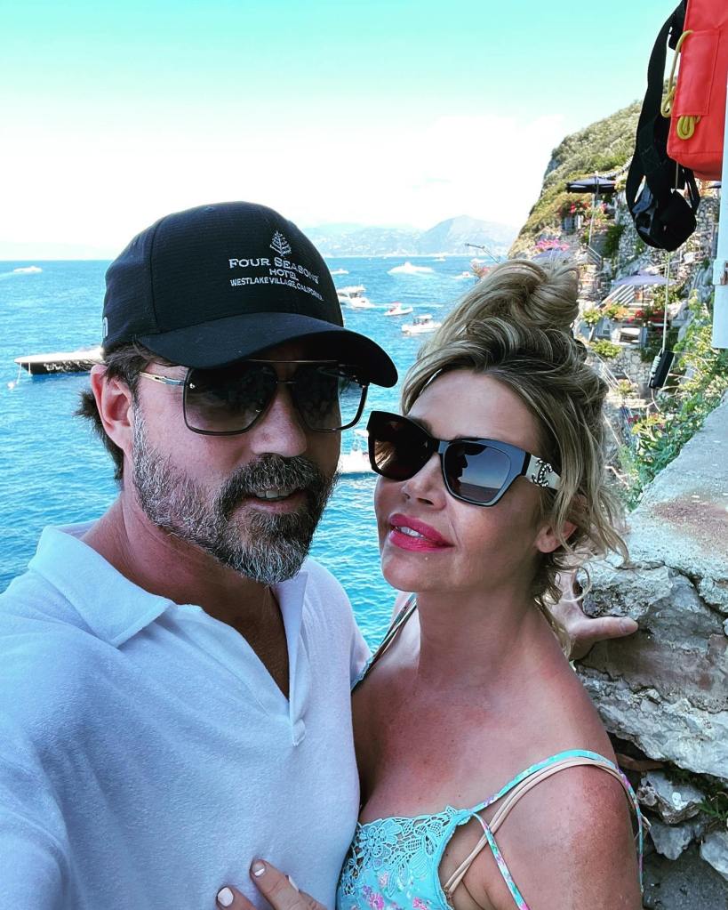 Denise Richards and Aaron Phypers posing for a selfie outdoors with the sea in the background.