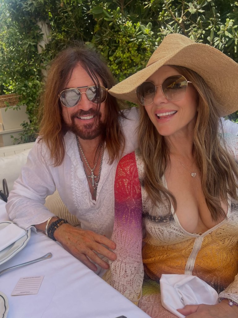 Elizabeth Hurley and a male companion smile together outdoors, both wearing sunglasses and summer attire.