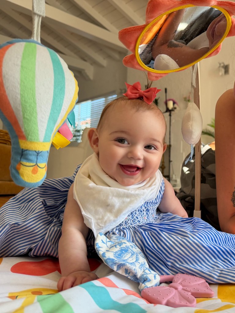 A smiling baby wearing a blue and white striped dress and a white bib, playing under a colorful baby gym with toys.