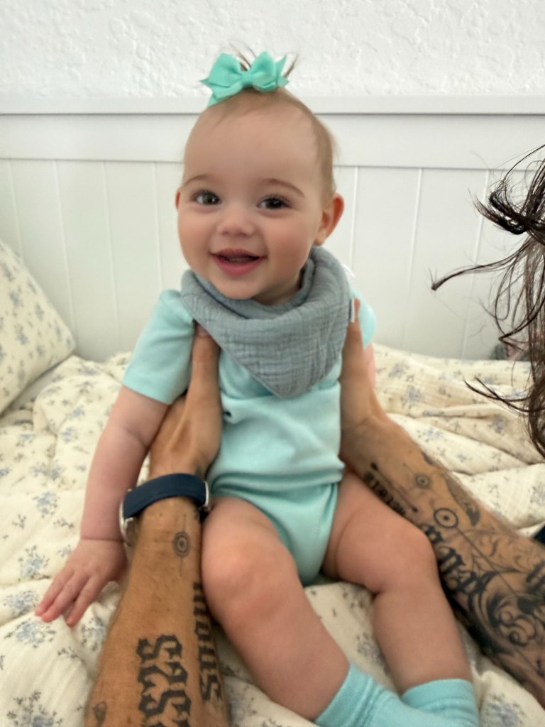Model Sahara Ray's daughter smiling in a light blue onesie and matching bow, held up by tattooed hands.
