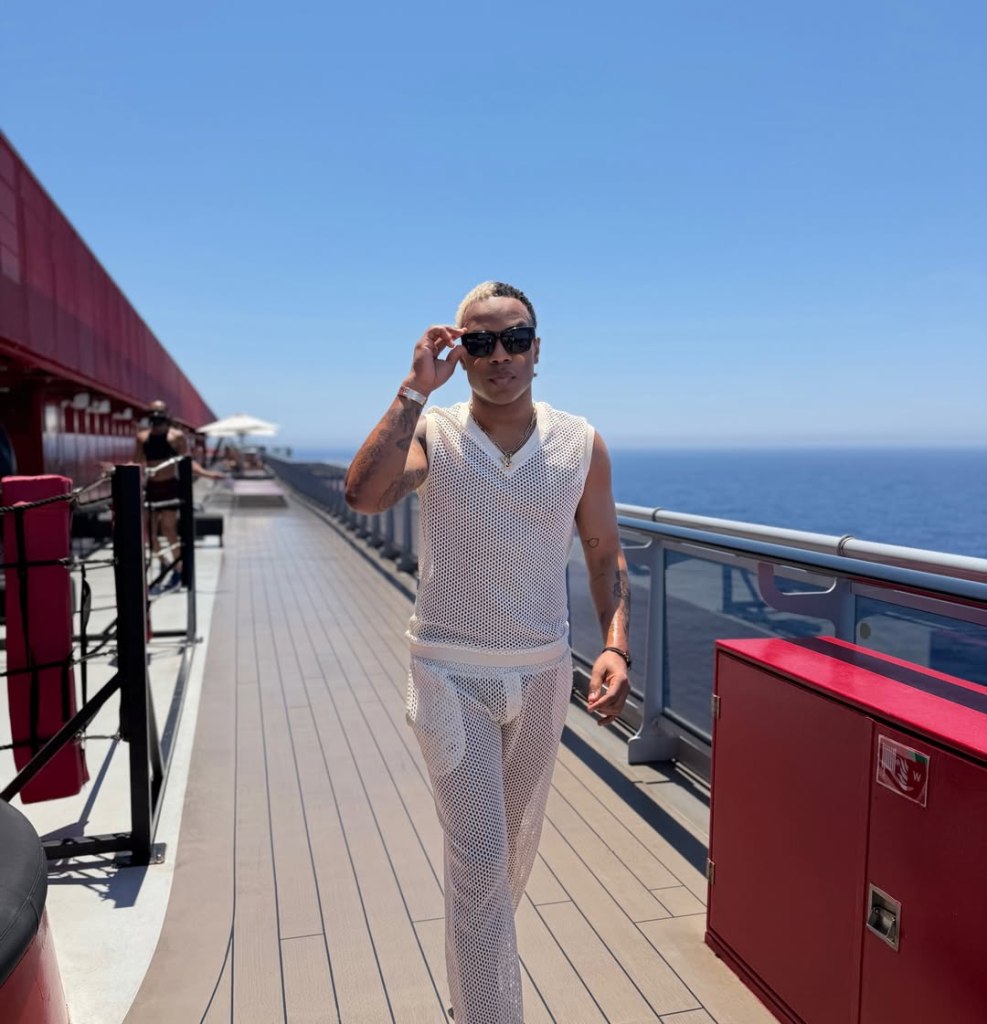 Todrick Hall walking on a cruise ship deck wearing a white mesh outfit and sunglasses.
