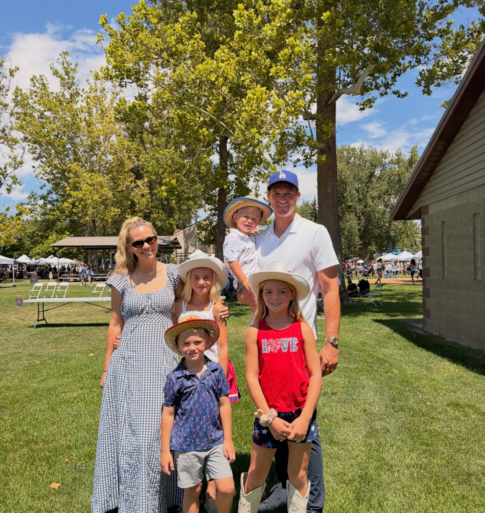 A family of six poses outside on a sunny day in a grassy park, with a mother, father, and four children wearing hats.